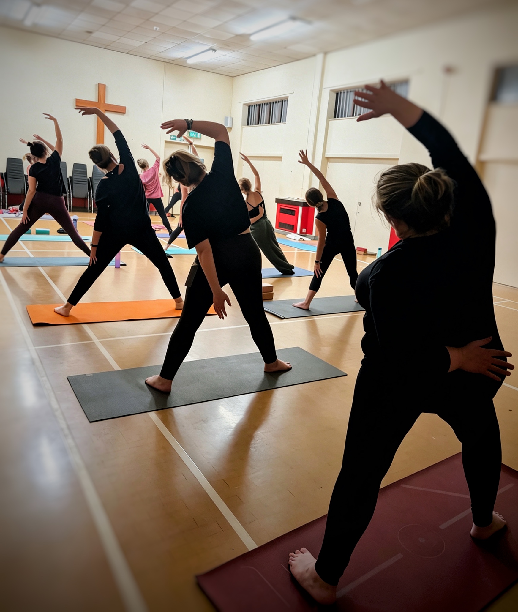Students holding a standing side bend across the wooden floor of Oakwood Church hall, arms reaching up and arching to the side; a large wooden cross is mounted on the far wall