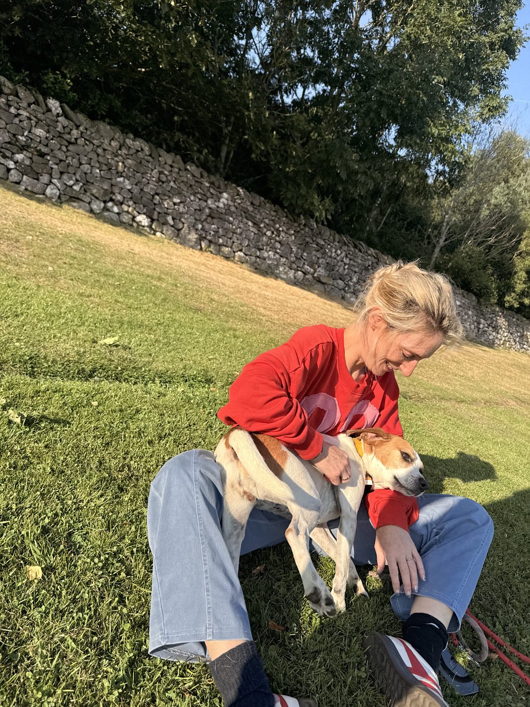 Jenny sitting on the grass in a red jumper, laughing with her dog