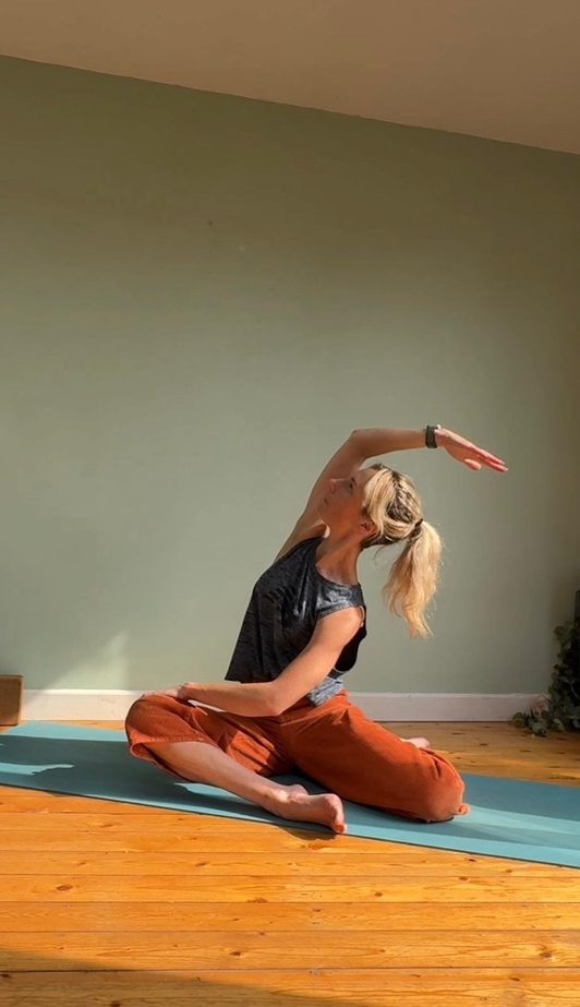Jenny seated in a cross-legged side bend, practising at home in front of a sage green wall